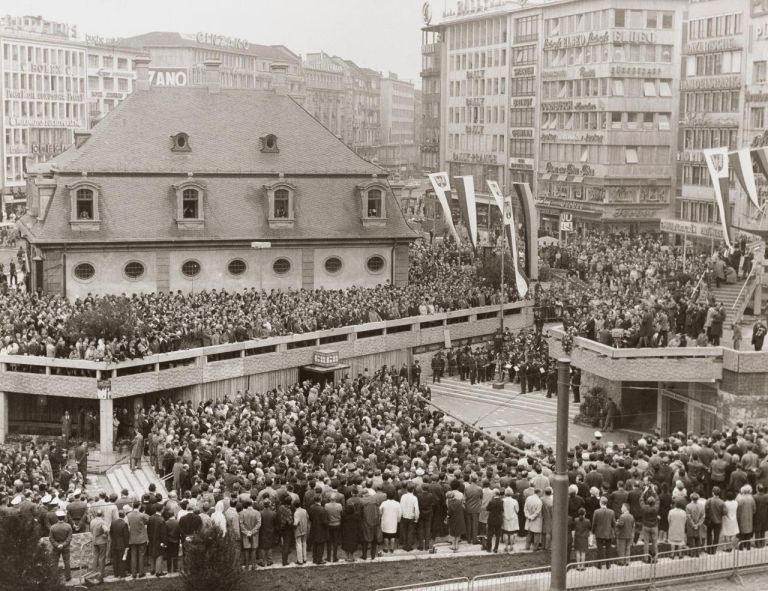 Schwarz-weiß Fotografie, auf der eine Menschmenge auf einem Platz mit einem Eingang in die B-Ebene vor Häusern steht